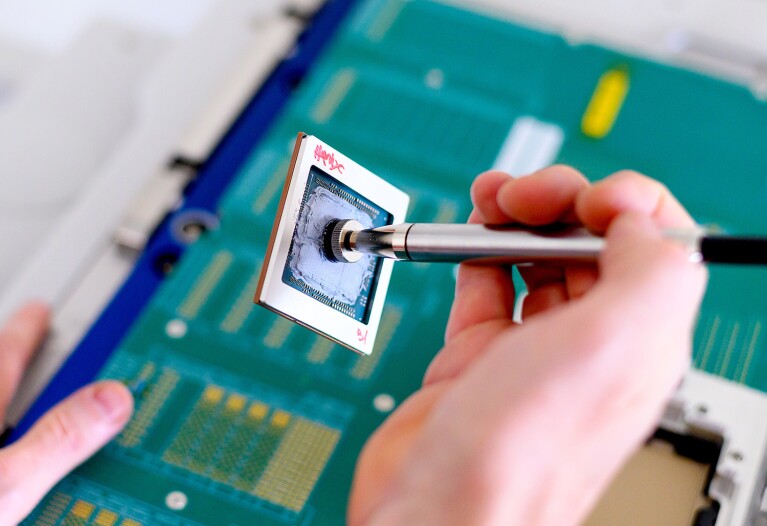 A person holds a metal pen with a small chip attached at the end. There is a computer board on the surface below.
