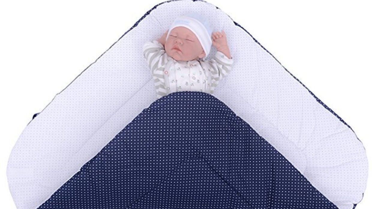 Un nourrisson dort, les bras levés, sur un matelas bleu à pois blancs. Il porte un pyjama blanc et gris et un bonnet blanc.