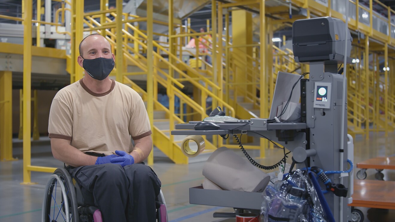 An Amazon Fulfillment Center employee wearing a black mask and protective blue gloves sits in a wheelchair next to a mobile computer desk.