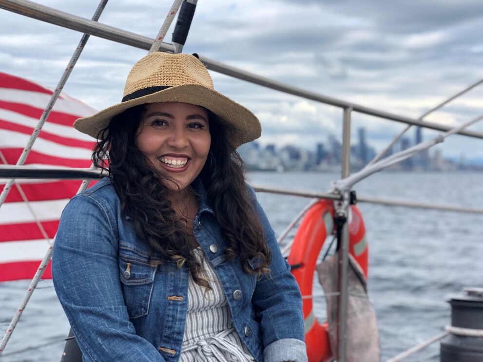 An image of a woman smiling for a photo while sitting on a sail boat. The American flag flies behind her on the boat.