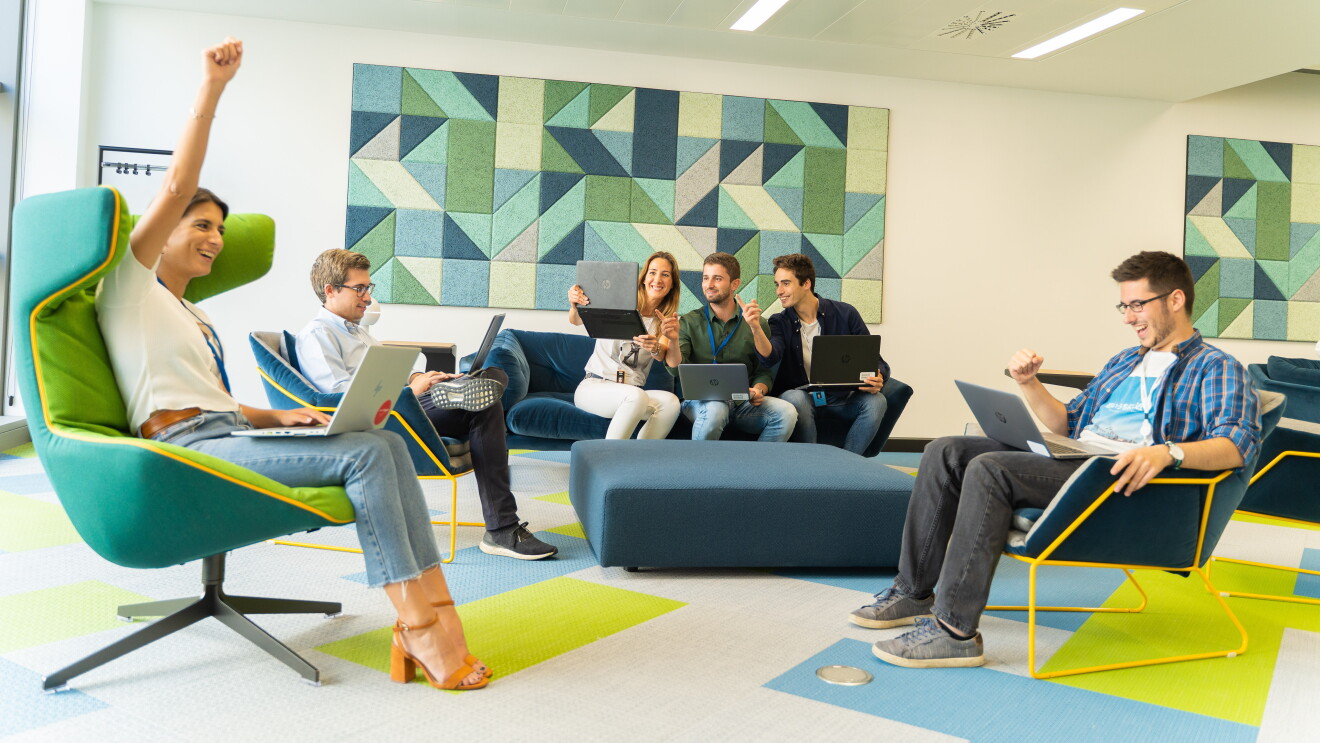 Parte del equipo que prepara el Prime Day se encuentra en una sala de las oficinas corporativas viendo cómo evoluciona el día. Tres de ellos están sentados en el sofá y tres más en sillones. Todos ellos tienen los ordenadores sobre las piernas. Los colores de las paredes y del suelo son amarillos y verdes y azules. 
