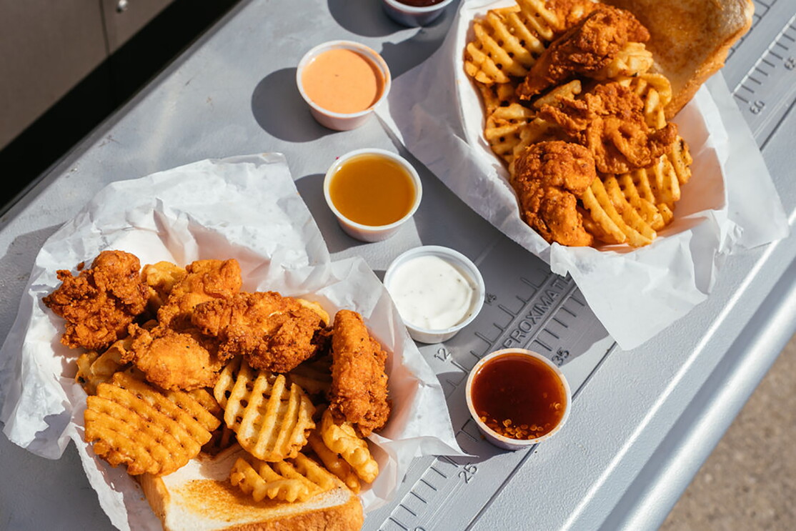 A basket of waffle fries, Texas toast, and chicken tenders, on ledge of a food truck window. To the right are four dipping sauces, and another basket of food. 