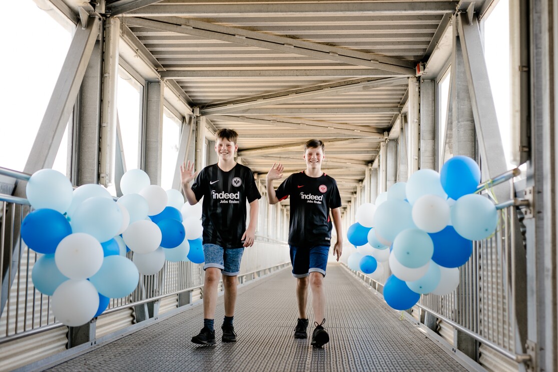 Zwei Jungen in schwarzen Fußballtrikos und Jeans-Shorts beim Betreten von Amazon in Bad Hersfeld. Rechts und links schmücken blaue und weiße Luftballons das Geländer.