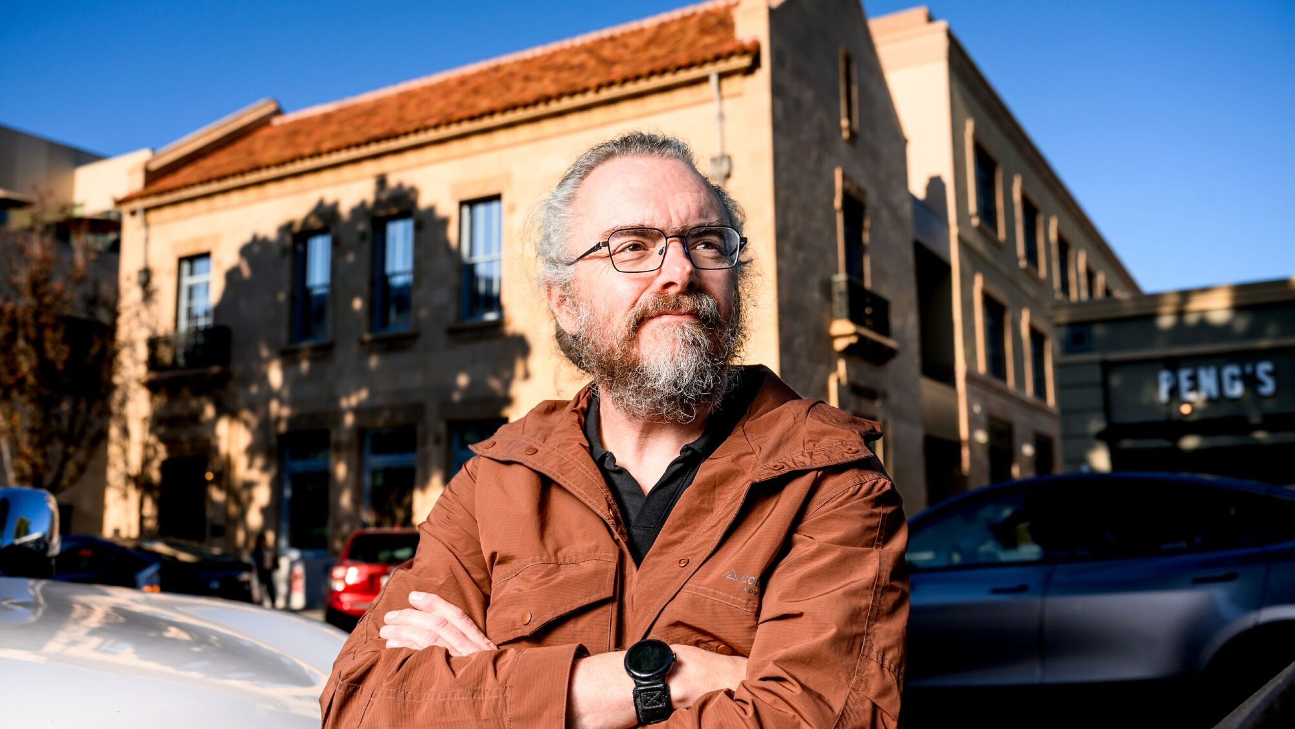 Paul Vixie in brown jacket standing confidently outside brick building.