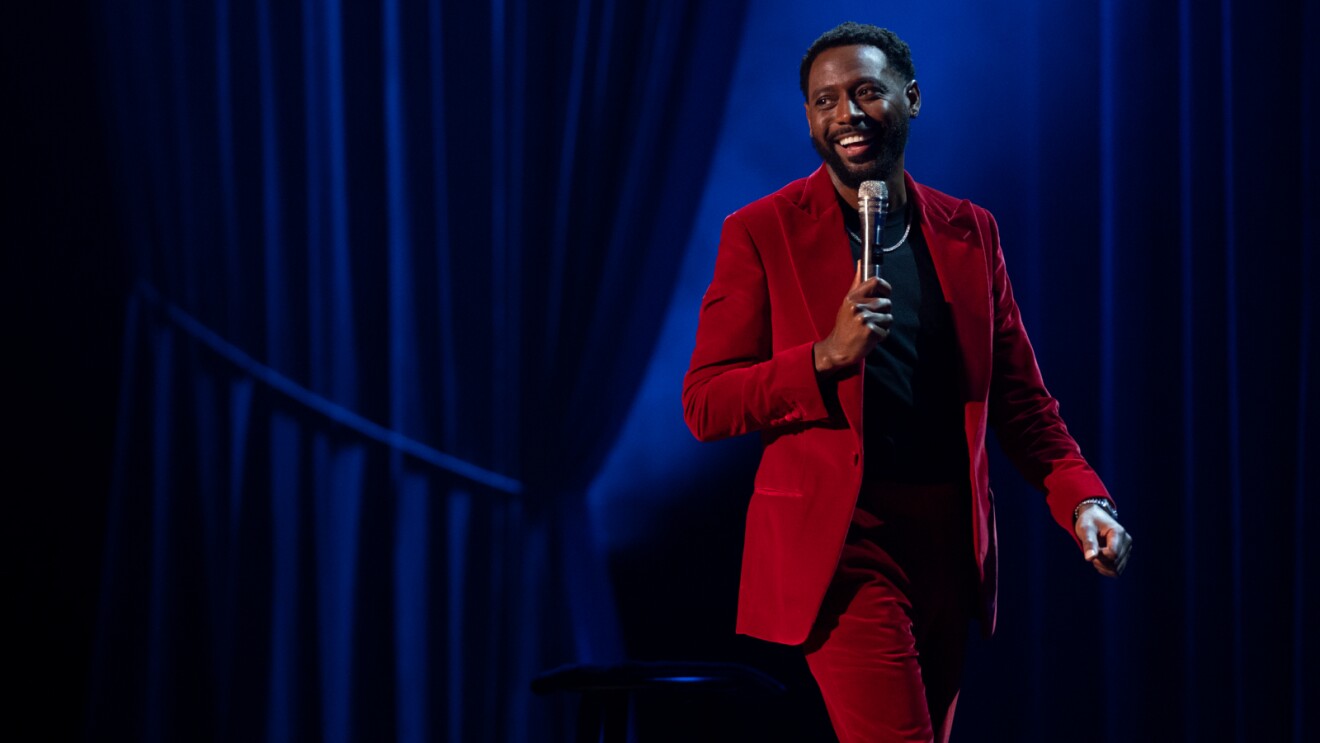 Kountry Wayne on stage giving a performance with a microphone in hand, wearing a red suit