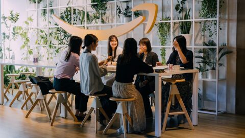 白い正方形で仕切られた棚の中に植物があり、その手前に大きな黄色いAmazonのスマイルロゴのレリーフがかけられている。その前に置かれたテーブルで6人の女性たちが話をしている