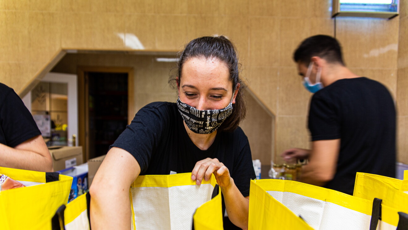 UNa voluntaria de Manos de Ayuda Social está preparando una bolsa de colida de color amarillo con las asas negreas. La volluntaria es joven, lleva una camiseta negra de manga corta, el pelo reocgido en una cola y una mascarilla negra con letras blancas que no se pueden leer. De fondo se ven dos voluntarios más de espaldas.  