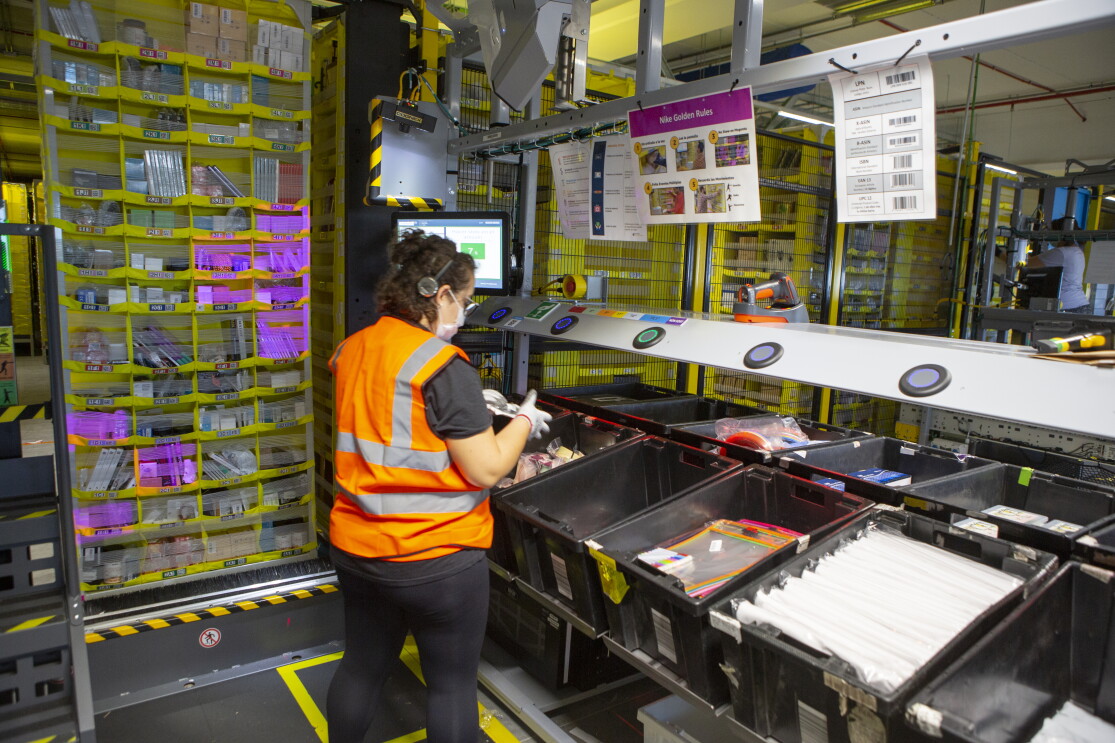 En un centro logístico de Amazon, de izquierda a derecha, aparece: una estantería amarilla llena de productos. Una trabajadora de Amazon de espaldas a la cámara. Lleva pantalón y camiseta negra, chaleco de color naranja y pelo rizado por el hombro. Está agarrando un producto con las dos manos. Se encuentra delante de una estación con cajas negras llenas de productos.  