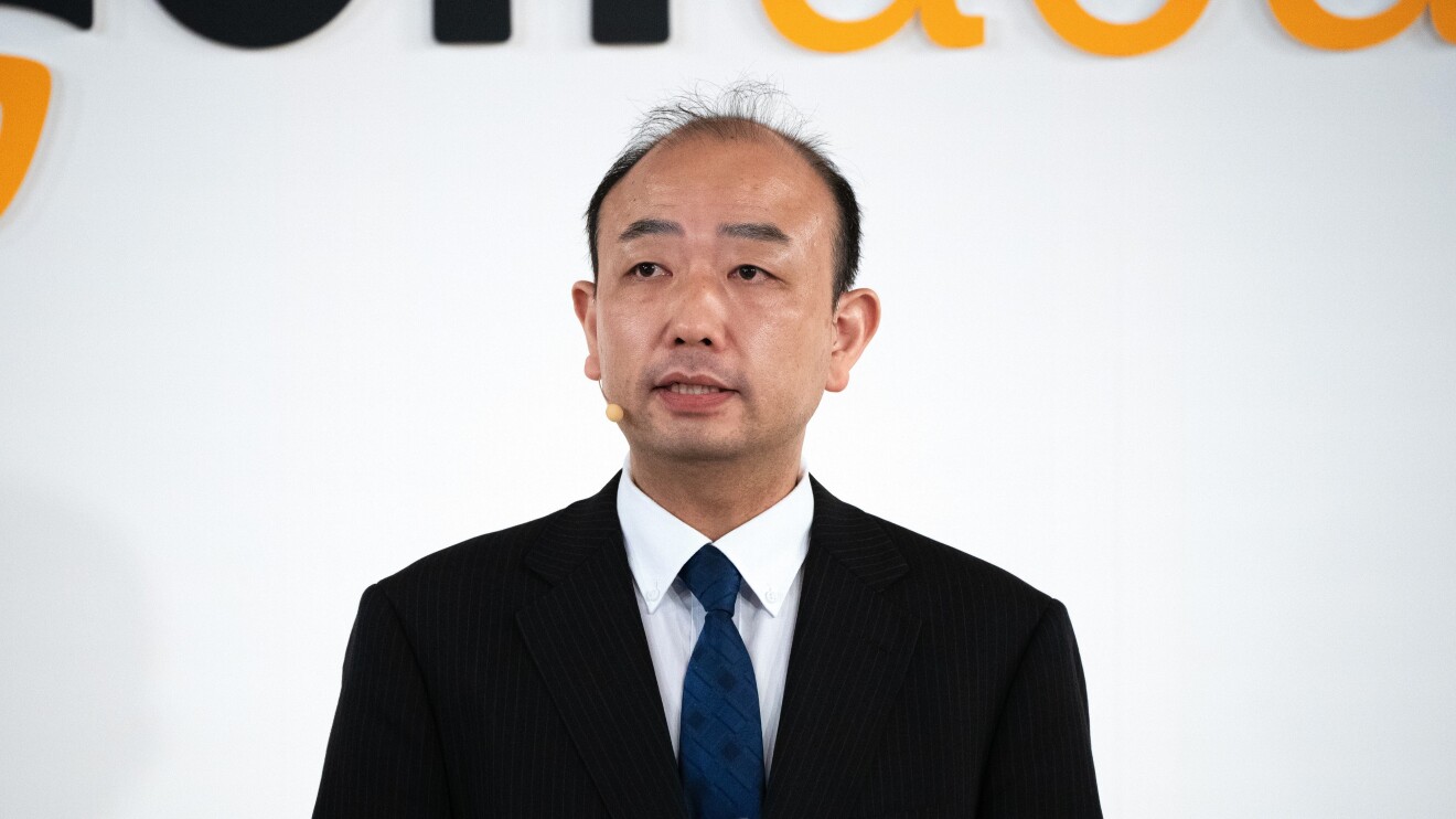 黒いジャケットに紺色のネクタイを着ている男性がヘッドセットをつけ話している(紺野博行氏)