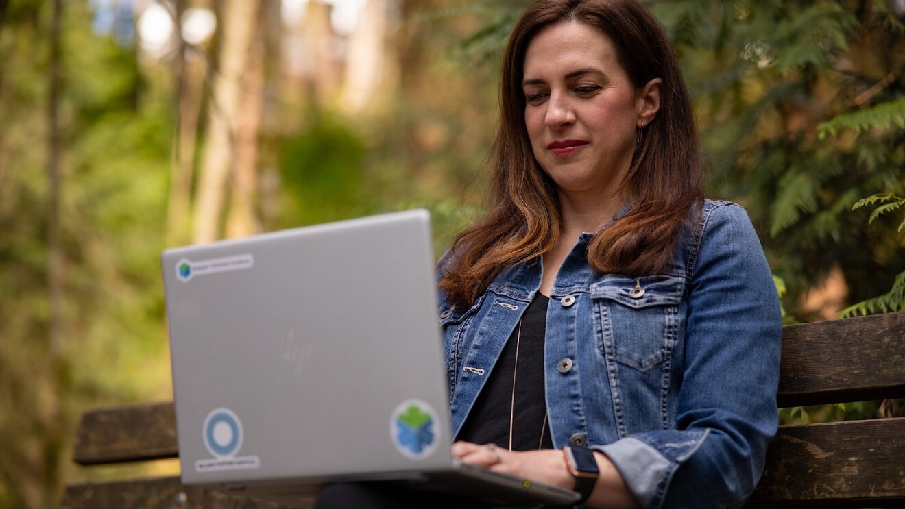 Ashely Rajagopal sits on a bench outside with vibrant greenery behind her. She is focused while she works on her laptop.
