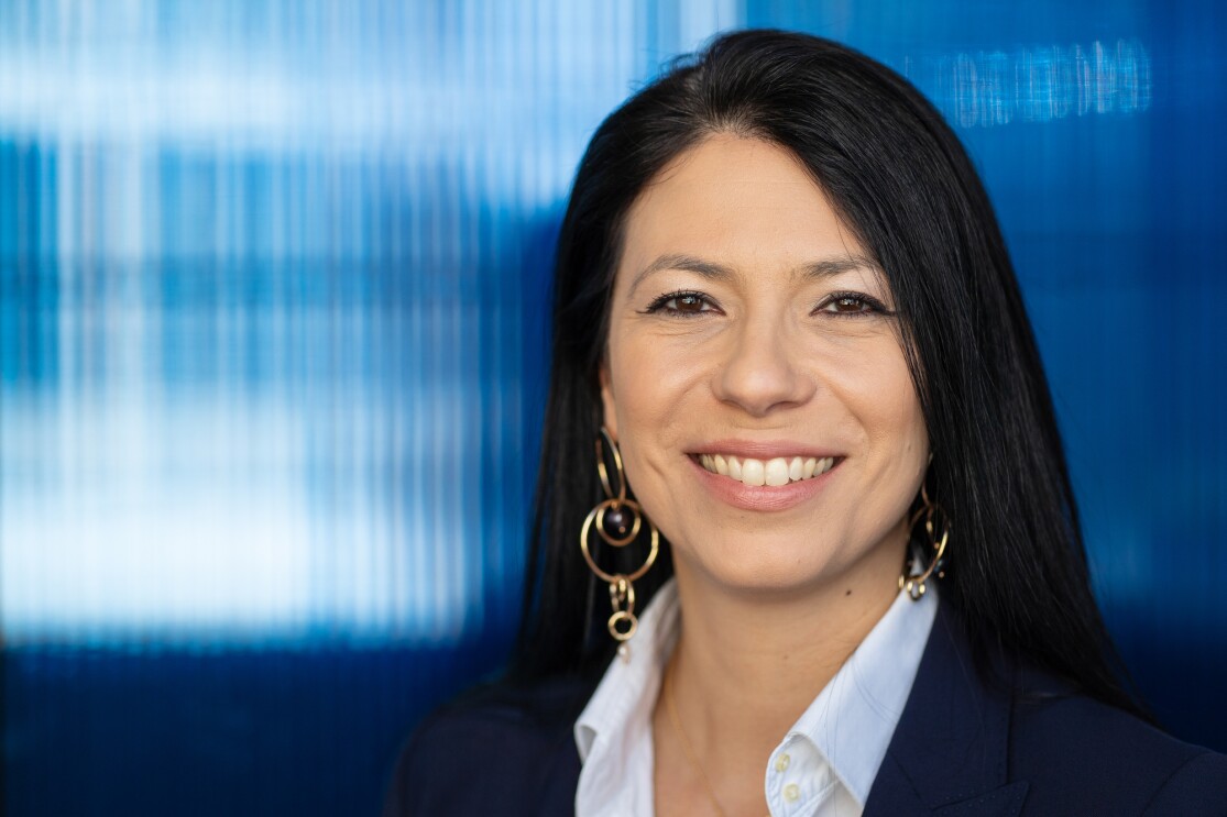 A woman wearing a blazer and white button down stands in front of a shaded glass wall. She wears gold drop earrings and smiles at the camera. 