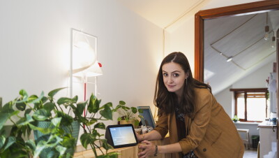 Elisa Reale, al centro della foto, è chinata verso un mobile. Guarda in camera, vicino alla mano destra ha un dispositivo Echo Show con lo schermo acceso. In primo piano, posata sullo stesso mobile, si vede una pianta in un vaso verde. Elisa indossa una giacca beige a righe bianche, ha i capelli lunghi e mossi. Sotto la giacca indossa una gonna a fiori colorata. 