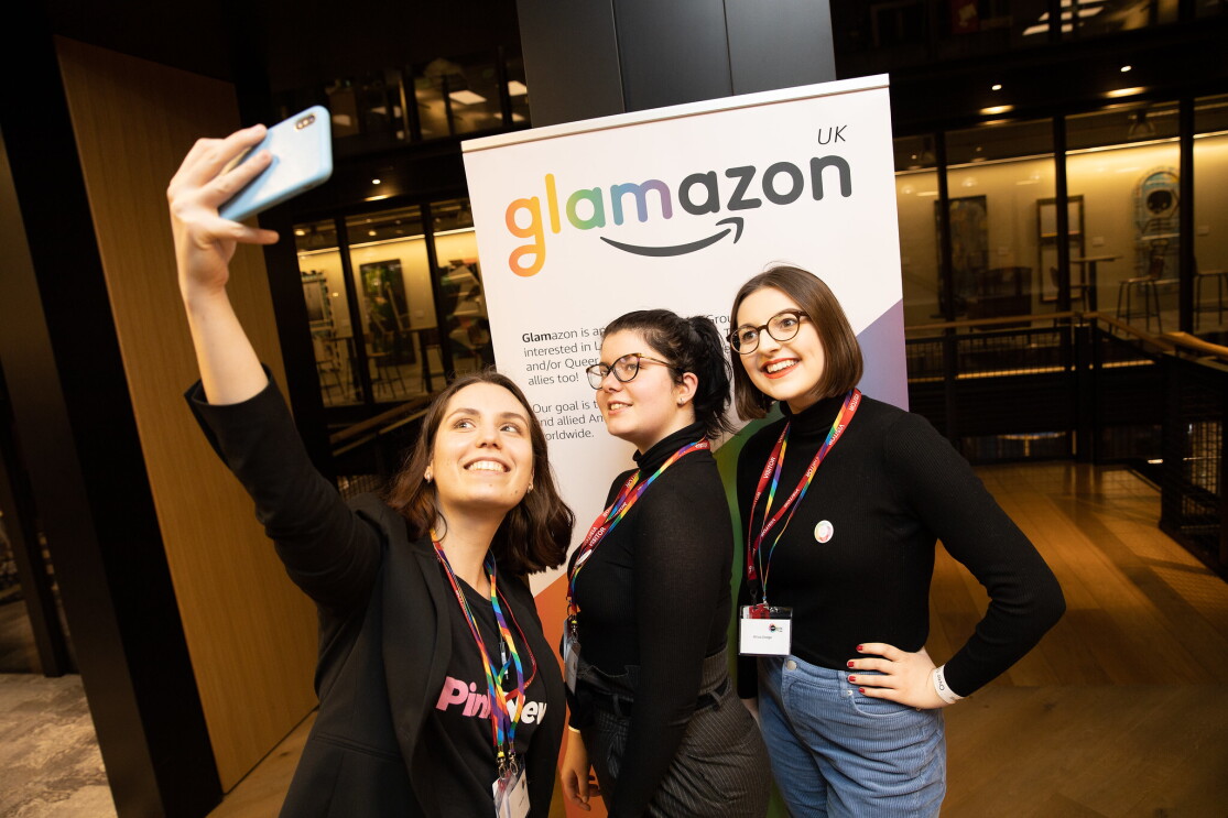 Two Amazon employees taking a selfie with a representative from PinkNews at the 2020 British LGBT+ Awards. They are standing in front of a glamazon banner. 