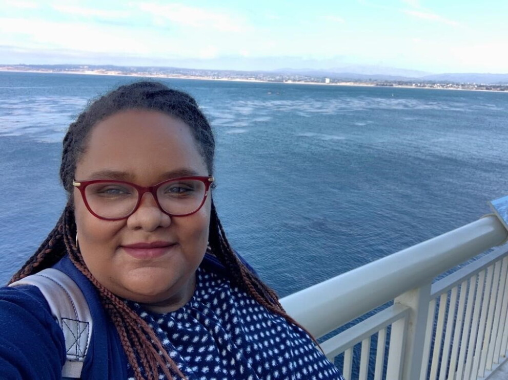 A woman smiles at the camera, with a smile on her face, in front of the ocean.