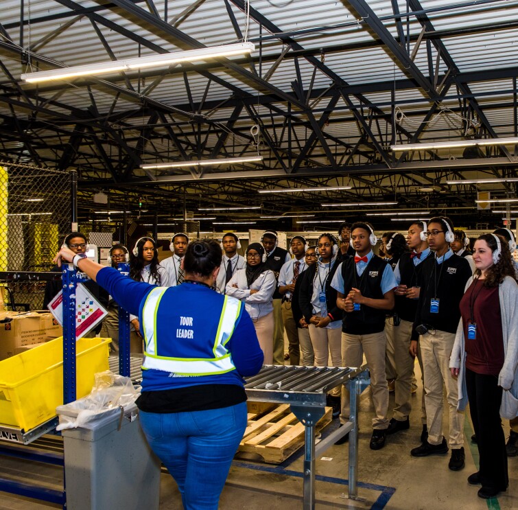 High school students wearing school uniforms and headphones stand in a group within an Amazon fulfillment center. They face an Amazon tour leader as she leads them through the facility.