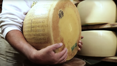 Primo piano di una forma di Parmiggiano Reggiano retta dalle mani di un uomo di cui non si vede il volto. Sullo sfondo, due scaffali con alcuni formaggi non stagionati.