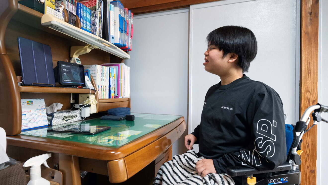 車椅子に座っている男性がデスクの上に置かれたタブレット（Echo Show 8）を見ている。タブレットの他には様々な本やホルダーなどがきちんと並んでいる。