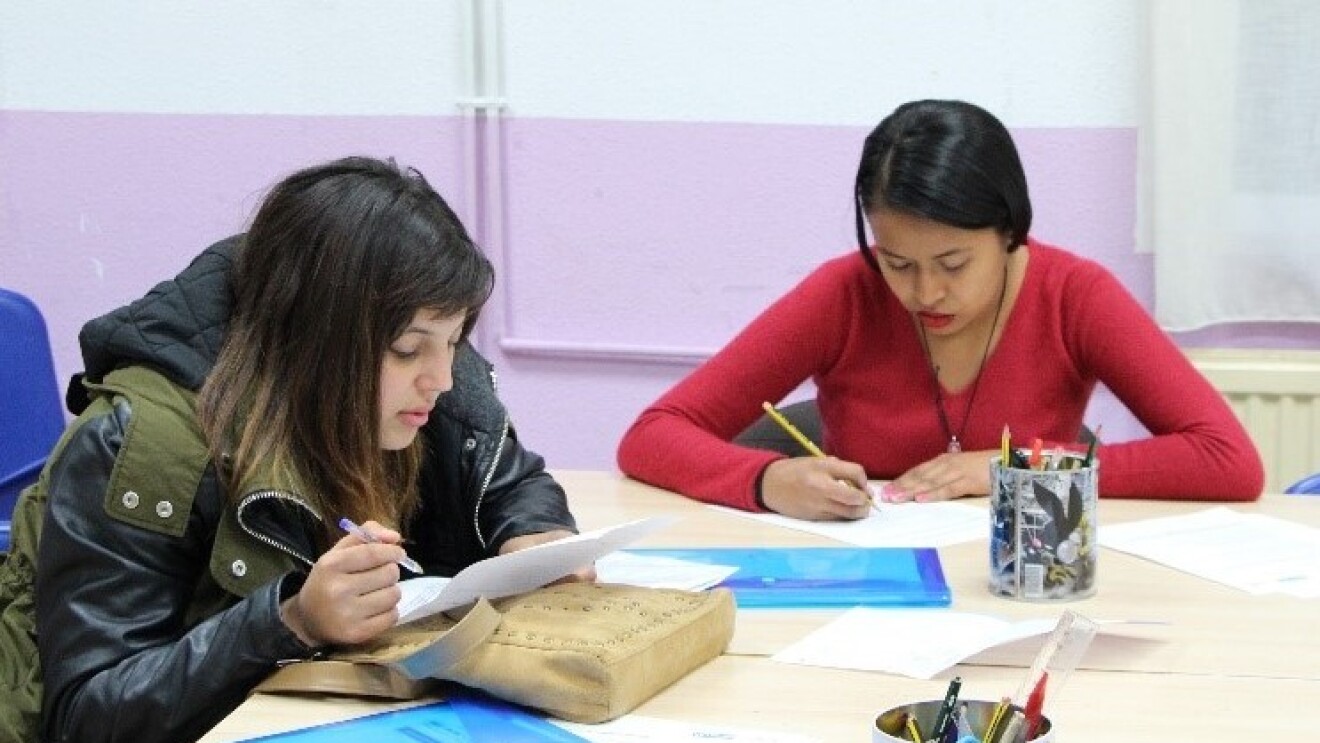 En una mesa blanca aparecen dos chicas estudiando. En la mesa hay hojas, dos carpetas azules y dos vasos con lápices y rotuladores. Una de las chicas lleva el abrigo puesto y lleva puesta una chaqueta negra y verde. Ella está sujetando una mano con un bolígrafo y en la otra tiene el papel. Está leyendo. El papel está sobre su bolso, que está encima de la mesa. Esta chica tiene el pelo oscuro y largo. La otra chica tiene un jersey rojo y está con un lápiz escribiendo en un papel.