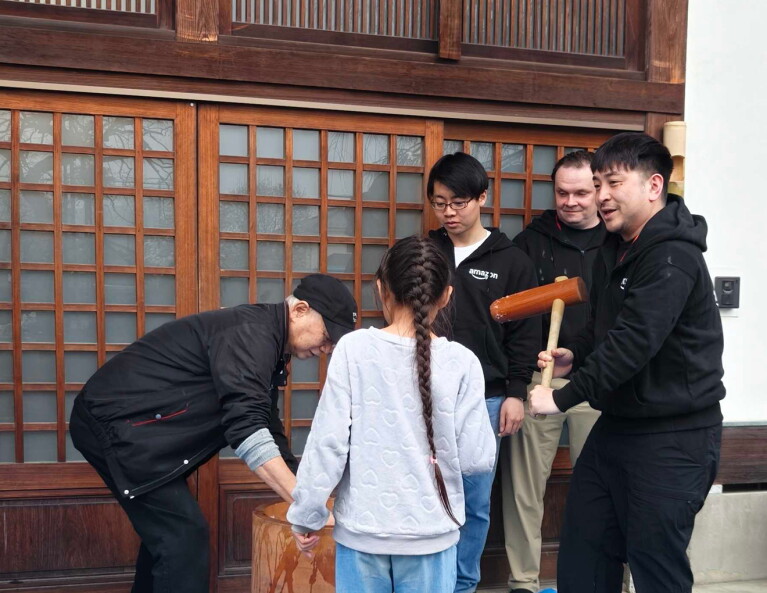 伝統的な日本家屋の玄関前で餅つきをする人々。左側の人物がうすにかがみこみ、右側の人物はきねを持っている。