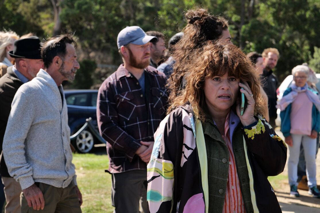 A curly haired woman wearing several layers of jacket is talking on her phone. Behind her are a small crowds seems to be interested in something. 