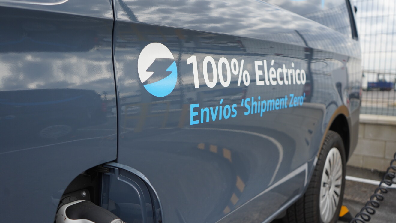 Una Mercedes Benz-Vans en un aparcamiento. Se ve el logo Prime de Amazon y el texto en la puerta 100% Eléctrico Envíos 'Shipment Zero'. En primer plano se ve un cargados de batería que está subministrando electricidad a la furgoneta de al lado que solo se ve un poco su parte trasera. 
