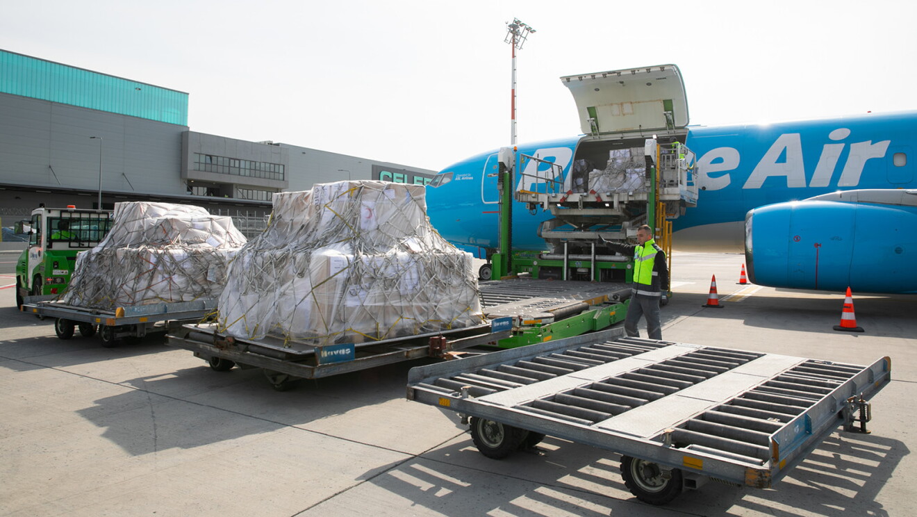 En la pista de un aeropuerto hay cuatro palés cargados con ayuda humanitaria. Los palés son de papel marrón, con pegatinas blancas y unas cuerdas. De fondo un avión de color azul con las letras en blanco Prime Aire. El cielo está nublado y es el atardecer. Los palés están arrastrados por una camioneta verde y blanca y el avión tiene una grúa por la que se está cargando la ayuda humanitaria. Hay un trabajador indicando dónde parar a la persona que arrastra, con una camioneta, los dos palés de ayuda humanitaria.  