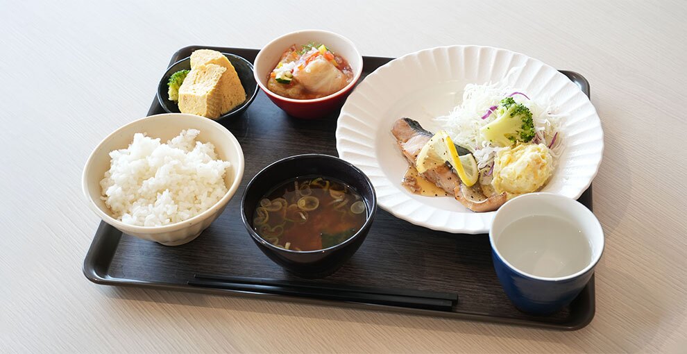 茶色の四角いトレーに、鮭のムニエル定食がのっている。白い皿にムニエルと千切りキャベツ、ポテトサラダ。その皿を囲むように左から黒い小鉢に卵焼き、赤い小鉢に和え物、白い茶碗に白飯、黒い椀に味噌汁。