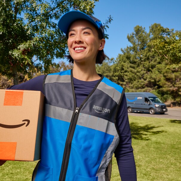 Woman wearing an Amazon delivery vest approaching a home with an Amazon box. Behind on the road is an Amazon Prime delivery van