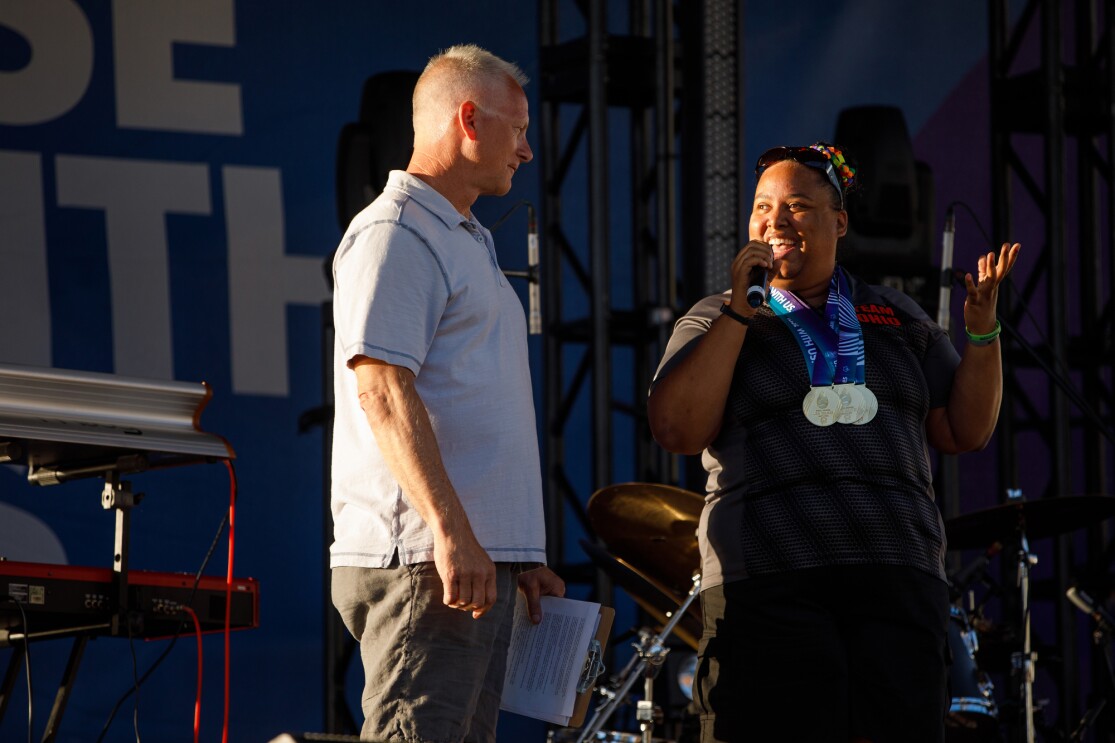 On stage, ESPN’s Kenny Mayne appears to be readying to take the microphone to speak during the USA Games closing ceremony. Next to him, Special Olympian Ali Riddick is speaking to Mayne and the audience, while wearing medals from the Special Olympics.