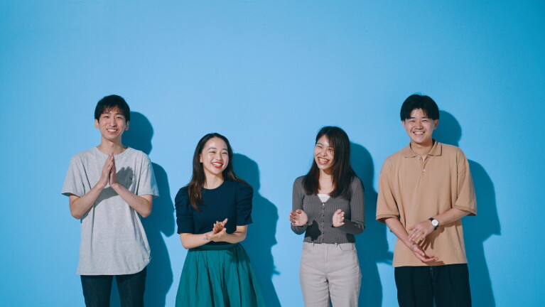 青い壁の前に男性二人と女性二人が笑顔で談笑している