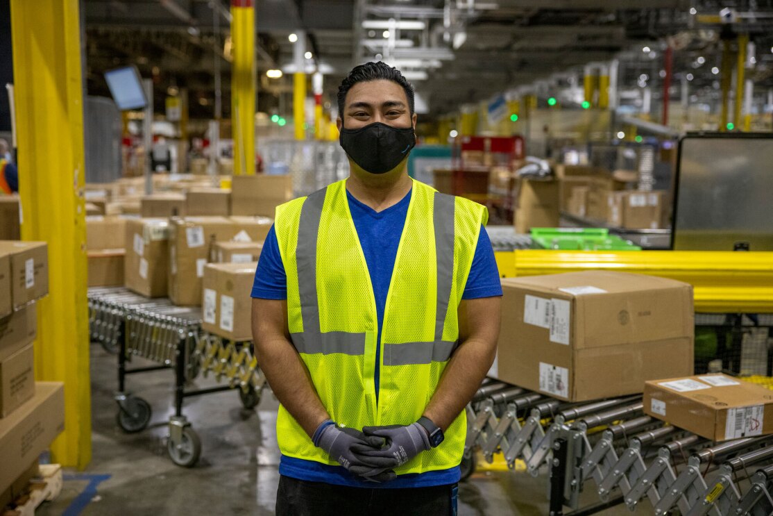 A male employee wearing a face mask and a yellow safety vest stands on the floor of an Amazon fulfillment center, looking at the camera. Boxes are on a conveyor belt behind him.