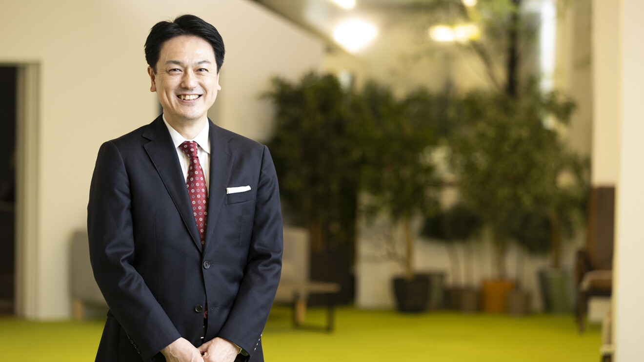 室内、笑顔で立つスーツ姿の男性。背景に複数の観葉植物が並んでいる。床はグリーンで、ソファが置かれているのが見える。