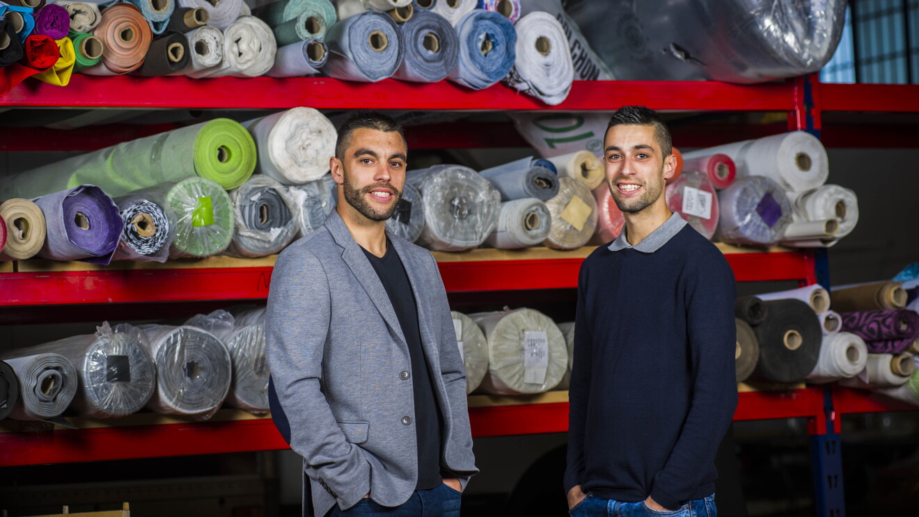 Los hermanos Javier y Daniel Mayorga con la marca SuenosZzz, los dos con las manos en los bolsillos, posan delante de las telas con las que tapizan sus productos. 