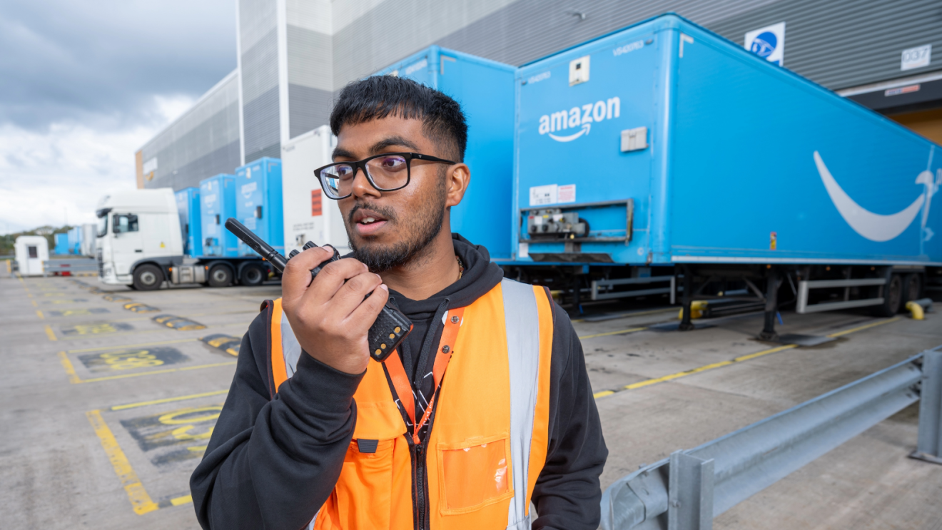 Fulfilment centre employee in a high-vis jacket talking into a walkee-talkee outside a FC, with Amazon lorries in the background
