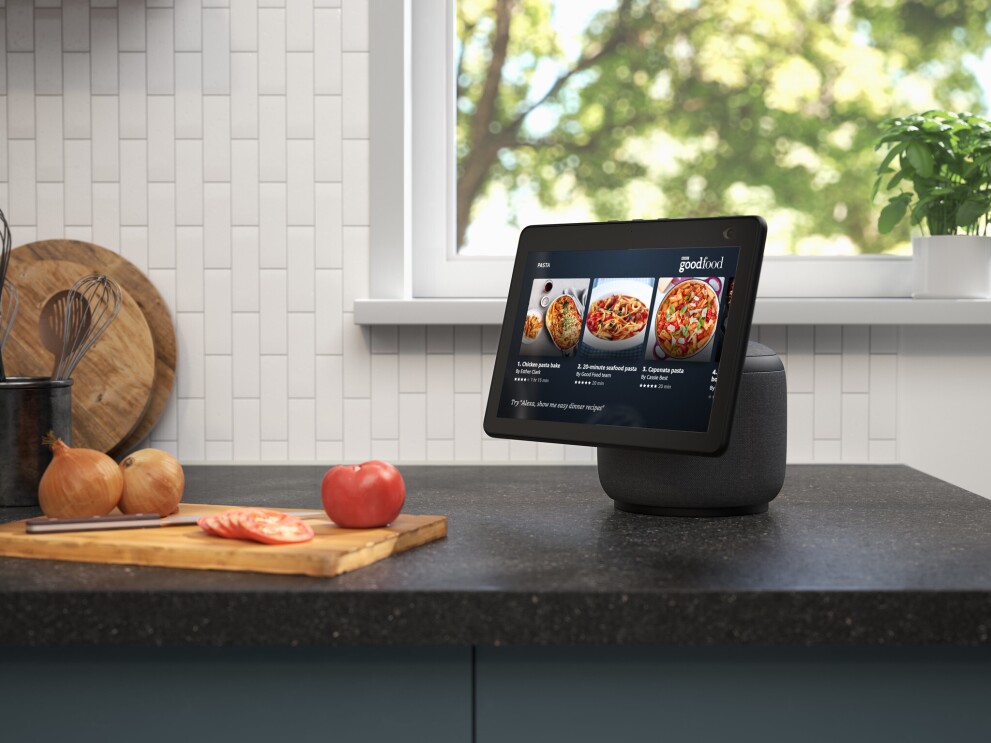 An image of the new Echo Show 10 on a kitchen counter. The device is showing recipes on BBC goodfood and next to the device there is a chopping board with tomatoes and onions. 