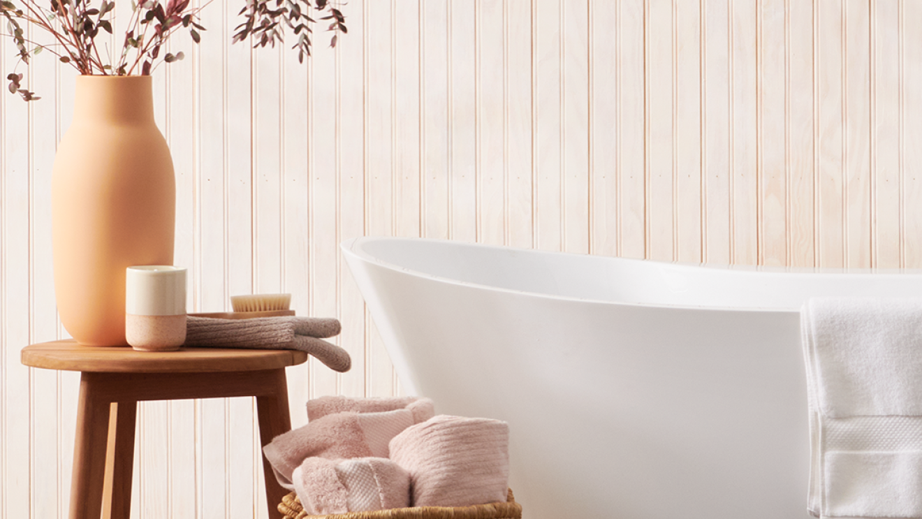 Foto de un baño al estilo bogegón. La pared es imitación madera con tablas verticales de color blanco roto. Una bañera de color blanco de las que no están pegadas a la pared. Con una toalla blanca apoyada. En el centro, un saco de mimbrte con tres toallas rosas sobresaliendo. Y a la izquierda un taburete con un vaso de color blanco y rosa, una toalla de color marrón, un esponja de baño estilo cepillo y un jarrón de barro con su color original con unas hojas secas de color morado oscuro.