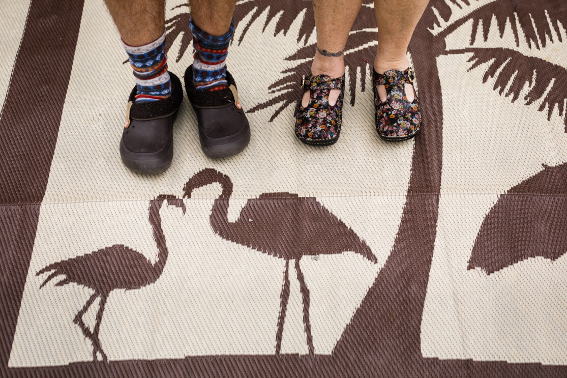 A man and woman photographed from the shins down. Both wear clogs. The man wears cozy socks. The woman goes sockless. They stand on an outdoor mat with images of flamingoes and a palm tree.