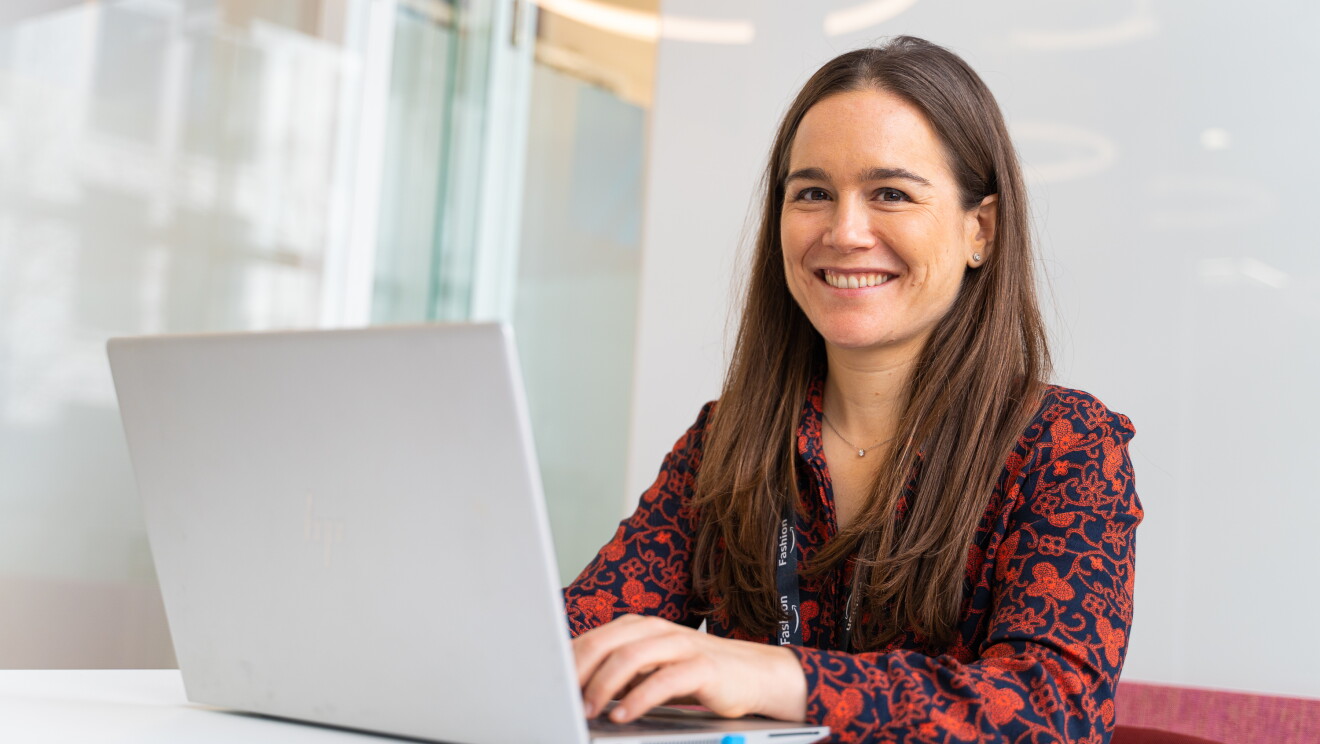 Lila Fernández es responsable de moda de los marketplace de Amazon en España e Italia y llegó a Amazon en 2016. Está sentada en una mesa de las oficinas de Amazon en Madrid. En una silla roja está trabajando con un ordenador. Lila mirando a cámara y sonriendo. Lleva un badge con una cadena de color negro. Una camisa azul con estampado de color rojo. Lleva pendientes y el pelo castaño, liso y largo. 