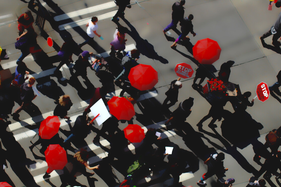 Protesters carrying red umbrellas and red signs that say STOP are photographed from above crossing an intersection. It is a photography piece of art.  