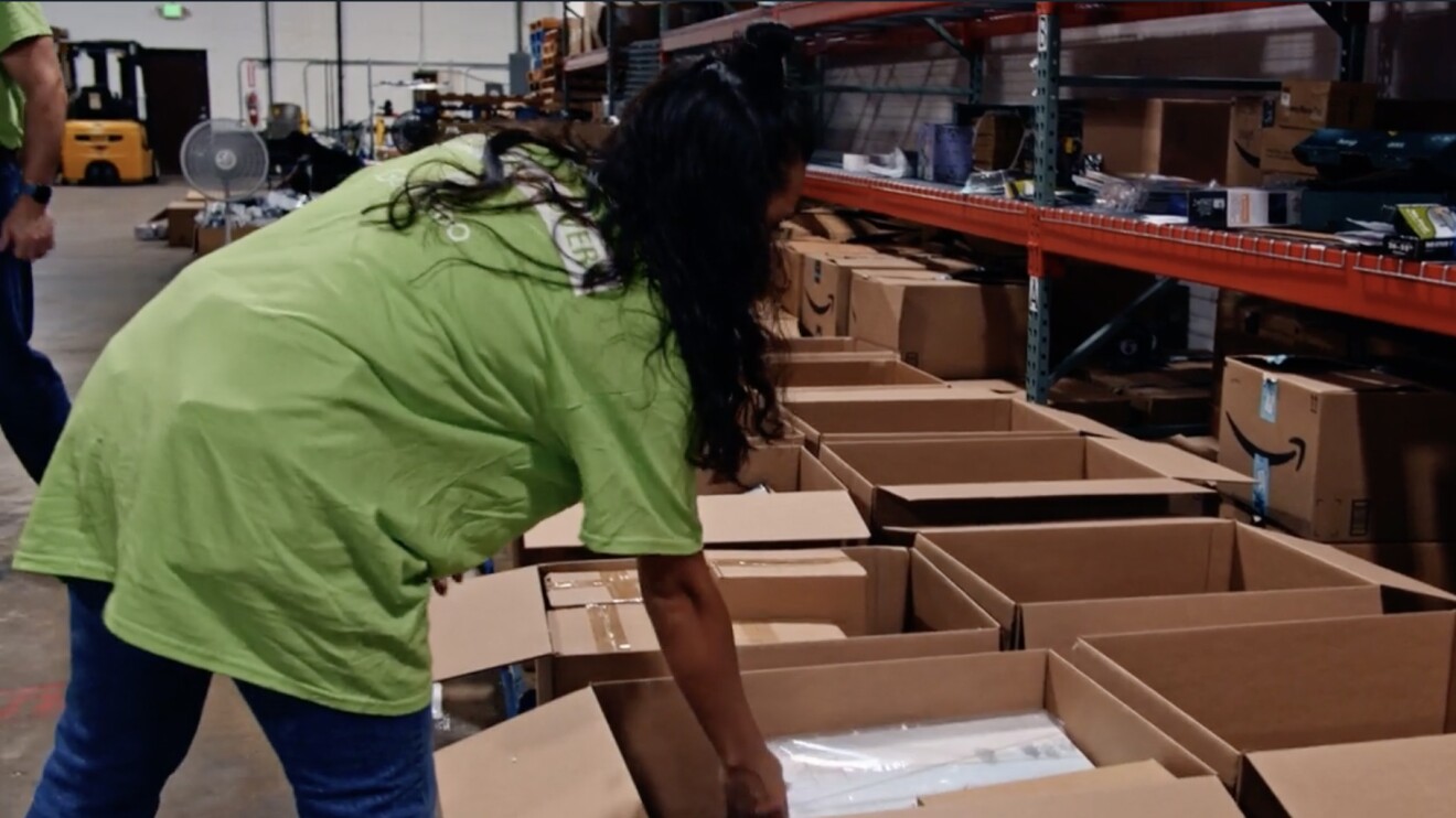 An image of a woman putting items inside Amazon boxes in a fulfillment center. 