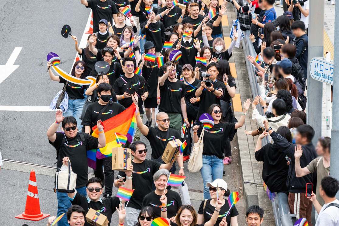 Amazonのロゴが入った黒いTシャツを着ている多数の人々が歩道にいる人たちに虹色の旗や手を振っている