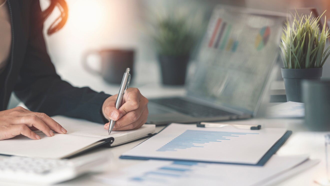 A woman writing at an office desk in a notepad, with a spreadsheets and her laptop also on the desk. 