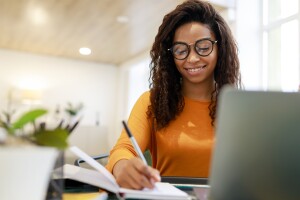 An image of a woman wearing an orange shirt and glasses. She is writing on a notepad with a laptop in front of her. 