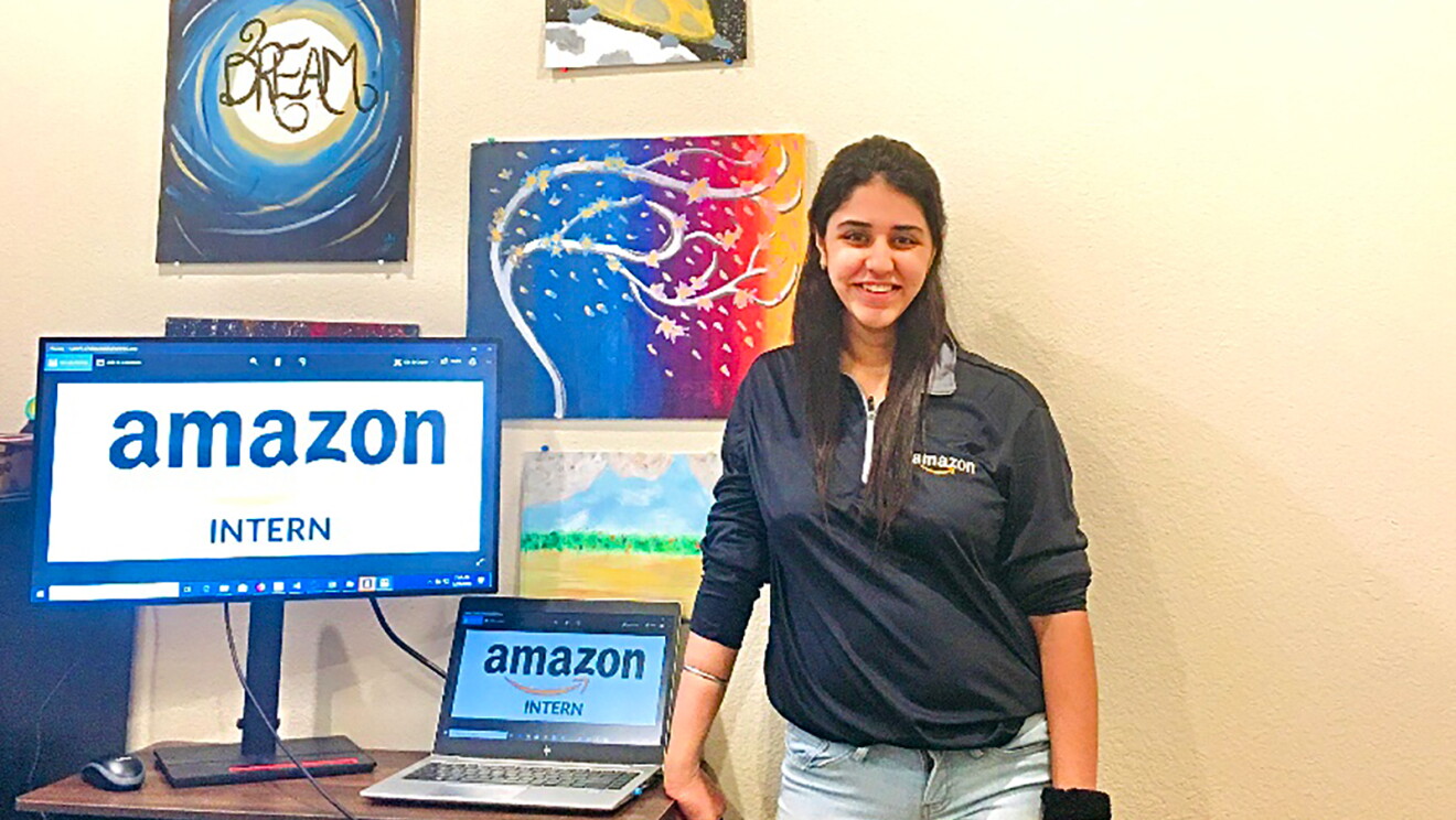 A woman stands next to a desk with a laptop and monitor displaying the words "Amazon intern."