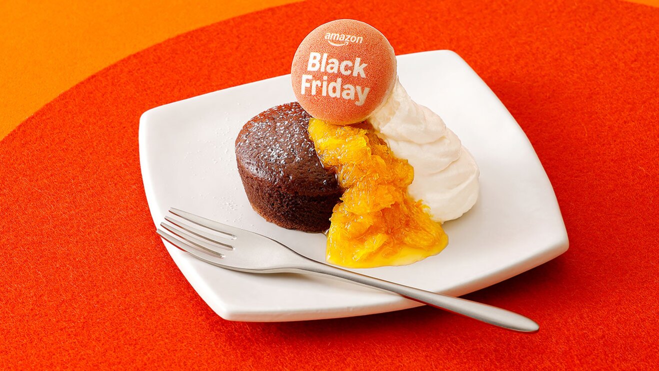 オレンジのテーブル、濃いオレンジのマットに、四角い白皿でガトーショコラを提供。マーマレードと生クリームを添え、上にマカロンがのっている。ケーキの手前側に銀のフォーク。