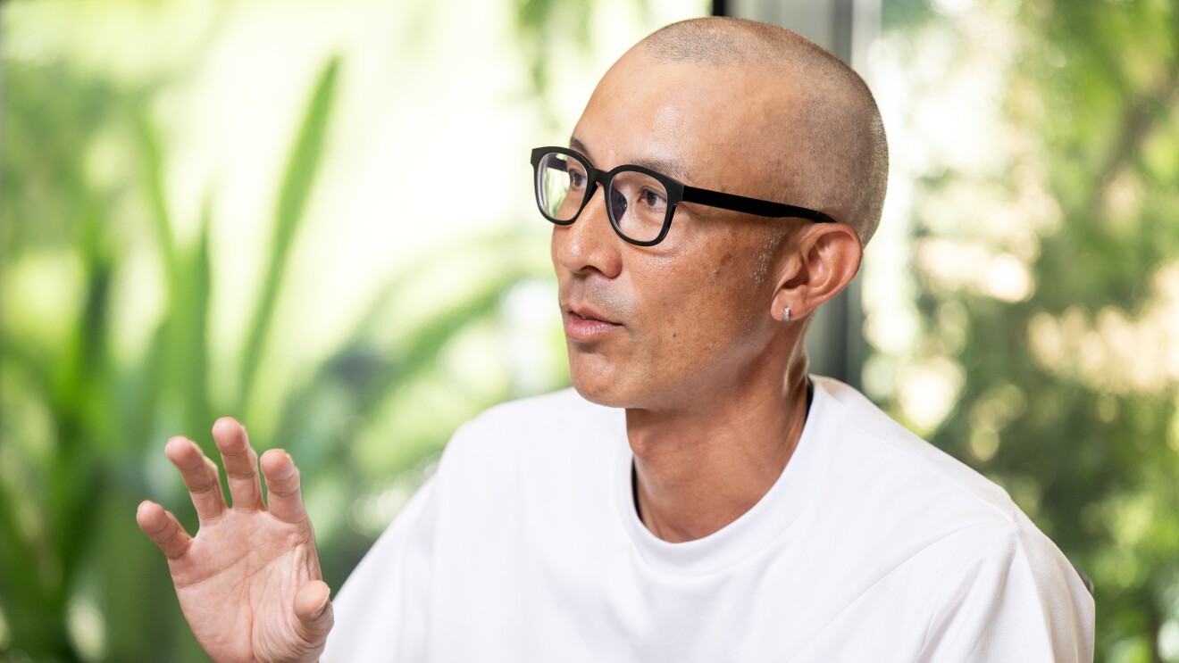 窓の外の緑を背景に話す人物のクローズアップ。黒ぶちの眼鏡をかけ、白いTシャツを着用している。右手を軽くあげてジェスチャーしている。