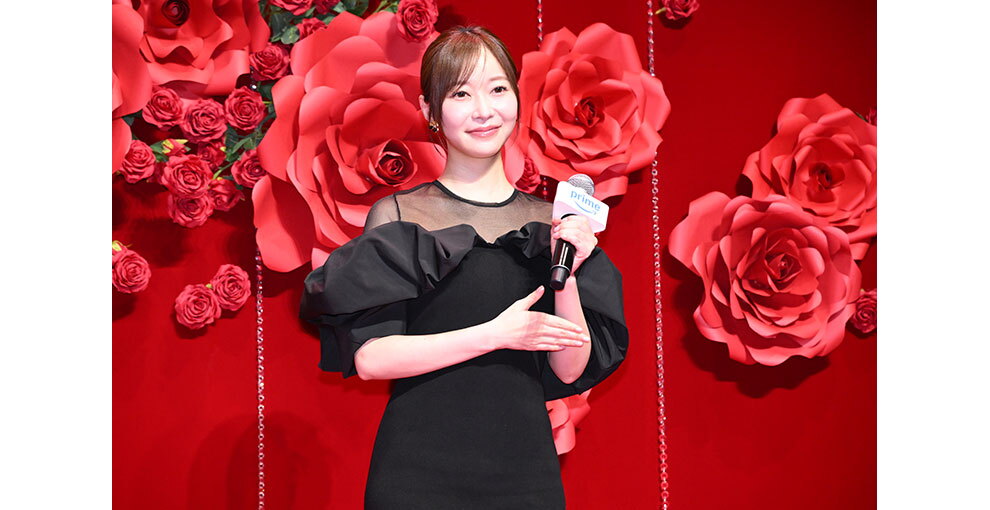 赤いバラの花の大きなオブジェと生花の赤いバラが飾られた赤い壁を背に、マイクを持ったドレス姿の女性。