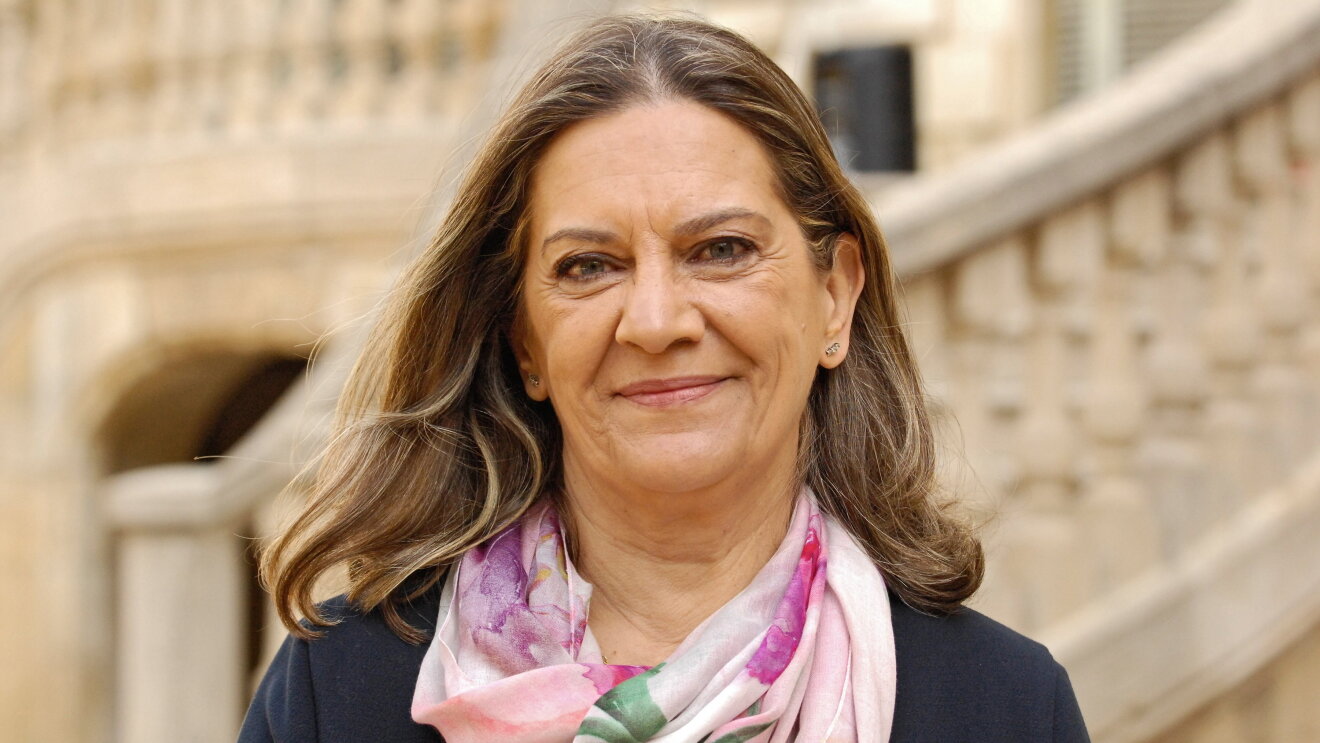 La escritora Mercedes Pinto con una chaqueta azul y un pañuelo a tonos rosa y morado. De ojos azules y pelo claro, mira a cámara sonriendo. El fondo de la foto es la escalinata del Palau Robert de Barcelona. 