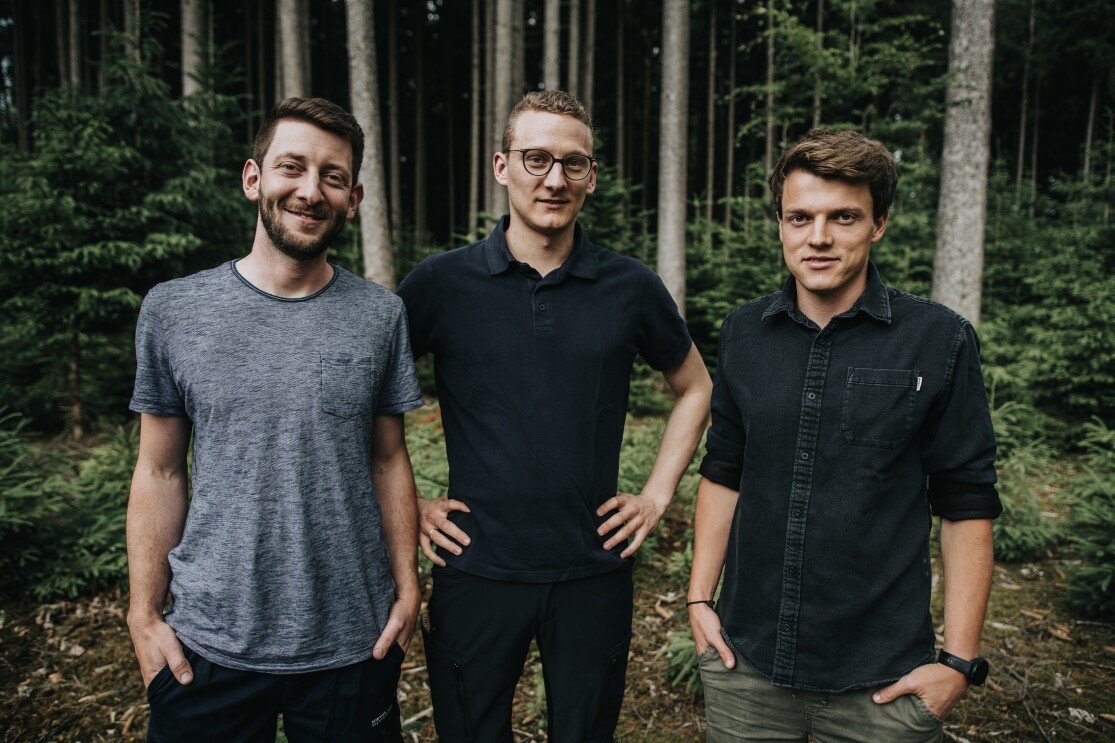 Drei Männer schauen lächelnd in die Kamera. Im Hintergrund sind Bäume in einem Wald zu erkennen.