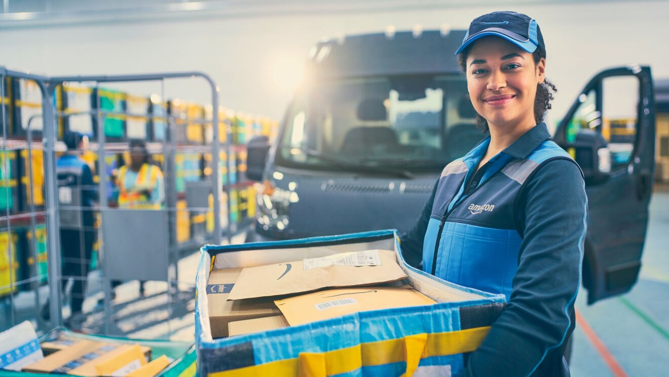 A photo of a delivery driver standing at an Amazon fulfillment center, holding a box that contains Amazon packages. A delivery van is behind them.